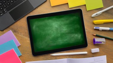 Tahta bir masanın üzerinde okul yönetimini taklit eden sağlam bir geçmişi olan bir tablet var. Okul malzemeleri etrafta..