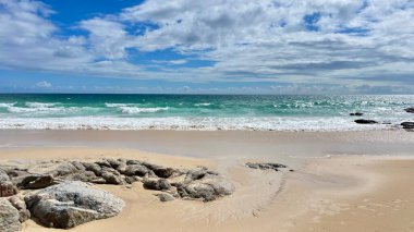 Yumuşak kumlu sahilli deniz burnu, ön planda kayalar ve turkuaz dalgalar. Sakin deniz ve bulutlu gökyüzü huzur ve rahatlama ortamı yaratır..
