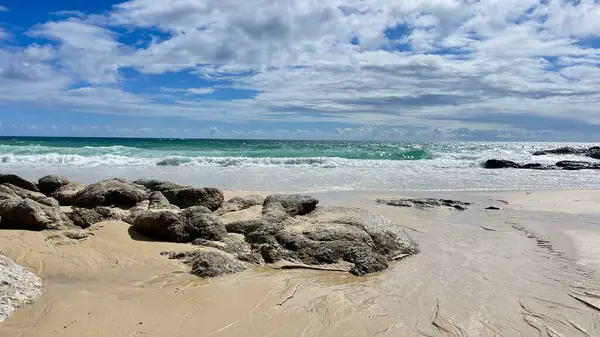 Ön planda kum ve kayalar olan terk edilmiş tropikal bir sahil. Turkuaz okyanus dalgaları, bulutlu parlak gökyüzü. Özgürlük, doğa, tatil ve sükunet kavramı.