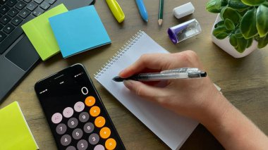 Bir adam defterine yazar, yanında hesap makinesi, çıkartma, kırtasiye ve dizüstü bilgisayarı olan akıllı bir telefon vardır. Planlama, çalışma ya da finansal hesaplamalar kavramı..
