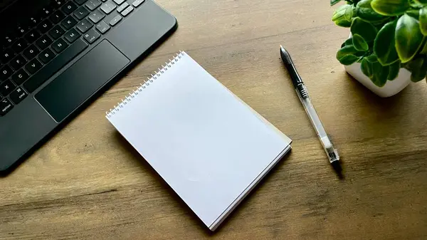Ahşap bir masanın üzerinde bir kalem ve yanında bir klavye ile birlikte bir defter var ve köşede bitkili bir çiçek var. Planlama, fikirler, öğrenme, notlar.