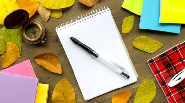 Beyaz sayfalı bir defter, bir kalem, bir pusula, kalemler ve parlak defterler sonbahar yapraklarıyla süslenmiş ahşap bir yüzey üzerinde yatıyor..