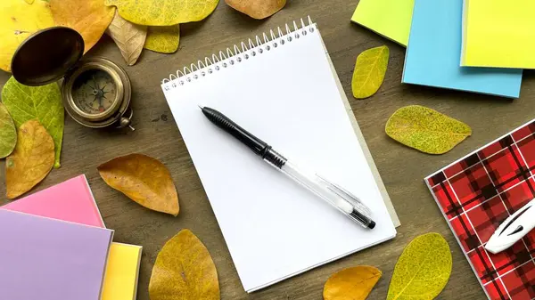 Beyaz sayfalı bir defter, bir kalem, bir pusula, kalemler ve parlak defterler sonbahar yapraklarıyla süslenmiş ahşap bir yüzey üzerinde yatıyor..