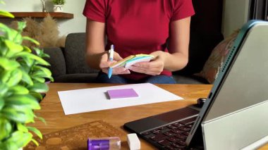 Genç bir kadın elinde tabletle masada otururken yapışkan bir nota notlar yazar. Uzaktan çalışmanın ya da evde internetten öğrenmenin rahat bir sahnesi.