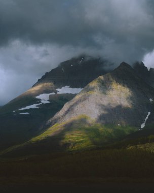 Kara bulutlarla kaplanmış karla kaplı bir dağın dramatik bir manzara fotoğrafı. Yeşil yamaçtaki bir yamacı aydınlatan bir güneş ışığı huzmesi.