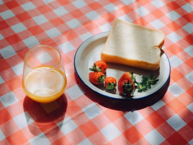 Yaz Kahvaltısı, bir bardak portakal suyu, bir tabak taze çilek ve bir dilim ekmek içeren basit ama davetkar bir kahvaltı, hepsi canlı kırmızı ve beyaz bir masa örtüsü üzerine dizilmiş.