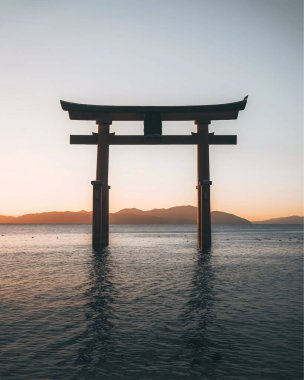 Görkemli bir Torii kapısı gölün sakin sularında tek başına duruyor, canlı bir günbatımı gökyüzüne karşı siluet..