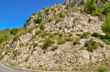 Kayalık arazisi, seyrek yeşilliği ve bulutsuz mavi gökyüzü olan engebeli bir dağ manzarası. Jeolojik, doğa ya da macera teması için uygun. Bir dağ yamacının doğal dokusunu vurgular.