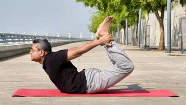 Dhanurasana 'da (Yay Poz) yoga yapan yaşlı bir adam, huzur dolu nehir kenarındaki gezinti alanında sağlık, esneklik ve iç huzura odaklanıyor..