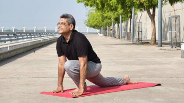 Ashwa Sanchalanasana 'yı (Binicilik Pozu) bir yoga minderinin üzerinde seslendiren kıdemli Hintli adam, sabah yogası sırasında nehir kenarındaki Ahmedabad' da esneklik, denge ve bilinçli nefes alma üzerine odaklanıyor. 