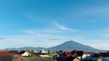 Güzel bir dağ manzarası sakin mavi bir gökyüzü altında dağınık bulutlarla, tuhaf bir köy üzerinde çerçevelenmiş. Doğal güzelliğin ve huzurlu yerleşim hayatının mükemmel bir tasviri..