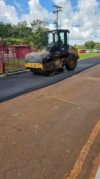 Siyah ve sarı bir asfalt makinesi güneşli bir günde yeni bir yol açıyor. Gökyüzü beyaz bulutlarla mavidir. Arka plandaki inşaat alanı ve bitki örtüsü şehir sahnesini tamamlar..