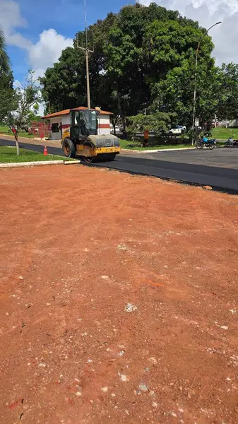 Bir sokakta yeni bir asfalt tabakasını sıkıştıran bir makine, arka planda yeşil ağaçlar ve parçalı bulutlu bir gökyüzü, inşaattaki bir günü resmediyor..