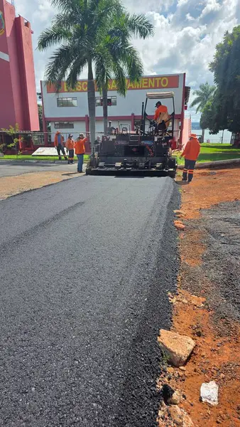 Turuncu üniformalı işçiler, açık ve güneşli bir gökyüzünün altında inşaat malzemeleri ve yeşilliklerle çevrili bir caddeyi asfaltlıyorlar..