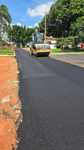 Arka planda yeşil ağaçlar ve binalarla çevrili, mavi gökyüzünün altına kırsal bir yol döşemeye çalışan bir kazıcı, altyapıdaki ilerlemeyi ve modernleşmeyi simgeliyor..