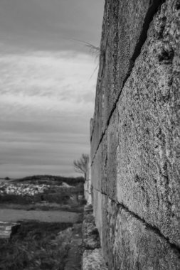 Bir tuğla duvarın gri ölçekli görüntüsü. Arka planda deniz kenarında büyük taştan bir duvar var.