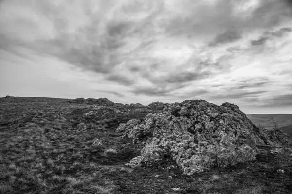 Bulutlu havada kayalık dağın gri tonlu görüntüsü.