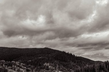 Dağların üzerinde siyah beyaz bulutlar ve arka planda orman var..