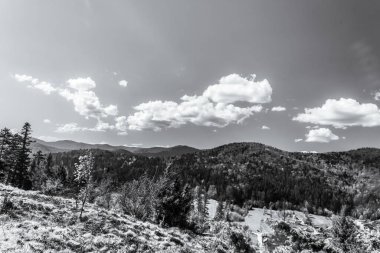 Arkaplanda, önplanda, bulutlu bir gökyüzünün altında bir dağ manzarası, bir yol ve ağaçlar.