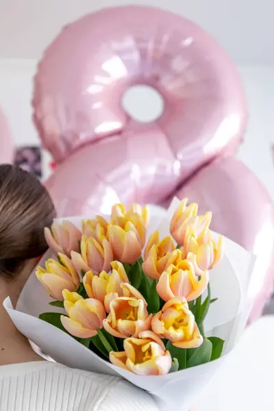 Genç bir kadın elinde pembe kenarlı taze sarı lalelerden oluşan bir buket tutuyor. Güzelce beyaz kağıda sarılmış..