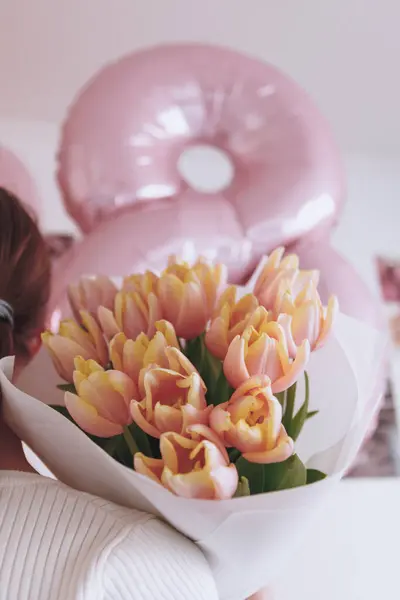 Genç bir kadın elinde pembe kenarlı taze sarı lalelerden oluşan bir buket tutuyor. Güzelce beyaz kağıda sarılmış..