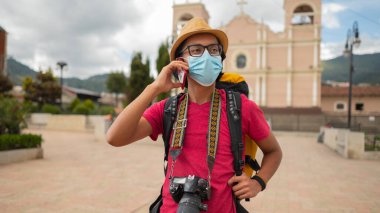 Portrait of a young man with a medical mask talking on a cell phone in front of a church.