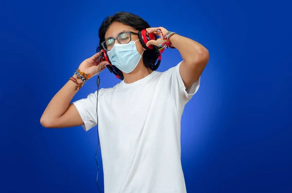Young man with headphones and glasses dancing and smiling using a medical mask on blue background