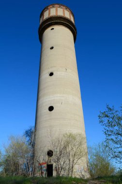 Altında grafiti olan eski, terk edilmiş bir beton su kulesi ağaçlarla çevrili açık mavi bir gökyüzüne karşı dimdik ayakta duruyor..