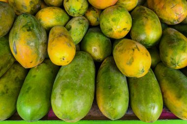 Geleneksel bir pazarda olgun ve olgunlaşmamış papayaların yakın çekimi. Tropikal meyveler canlı sarı-yeşil renkler ve doğal kusurlar sergiliyor, tazelik ve orijinalliği vurguluyor..