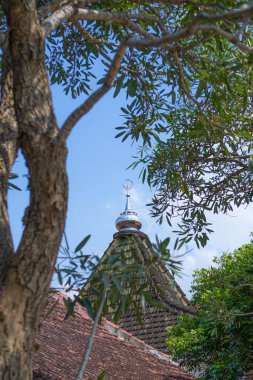 Geleneksel cami çatısı gümüş İslami ay hilal finali ile kısmen tropikal ortamda açık mavi gökyüzü altında yapraklı ağaç dalları tarafından çerçevelenmiş..
