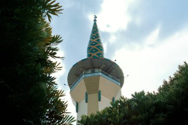 Bir cami kubbesinin düşük açılı çekimi. Tepesinde benzersiz geometrik desenler var. Canlı tasarım, yemyeşil, İslami mimariyi sembolize eden bulutlu gökyüzü ile çelişiyor..