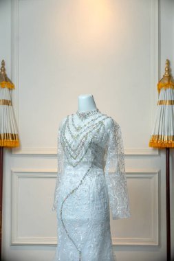 Karmaşık dantel kolları, parlak kristal boncukları ve detaylı nakışları olan zarif beyaz gelinlik resmi bir tören ortamında bir mankenin üzerinde sergileniyor..