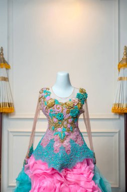 Altın, turkuaz ve pembe dantelli karmaşık çiçek işlemeli zarif bir balo elbisesinin yakın çekimi. Voluminous fırfırlı kumaşla eşleştirilmiş, bir mankenin üzerinde sergileniyor..