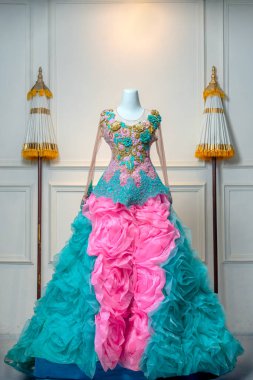 Zarif ve canlı bir balo elbisesi. Karmaşık çiçekli nakış ve pembe ve turkuazlı dramatik katmanlar. Klasik bir iç dekorda bir mankenin üzerinde sergileniyor..