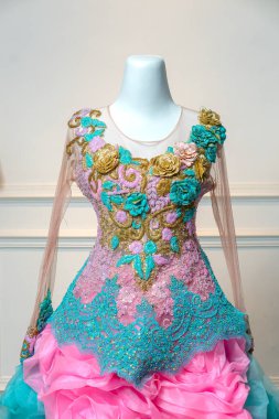 Saf uzun kollu zarif balo elbisesi, süslü altın, turkuaz ve pembe çiçekli nakış, ve katmanlı fırfırlı etek, klasik bir iç dekorda bir mankenin üzerinde sergilenir..