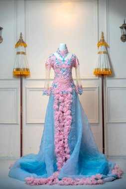 Zarif pembe ve mavi balo elbisesi, karmaşık dantel işlemeli, katıksız kollu, ve çağlayan çiçekli fırfırlar, geleneksel dekoratif unsurlarla geleneksel bir ortamda bir mankende sergileniyor..