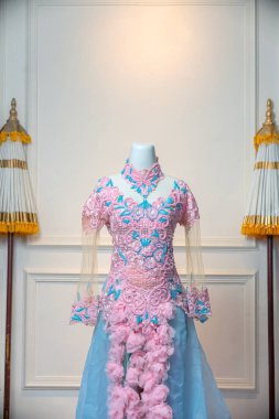 Zarif pembe ve mavi elbise, karmaşık çiçekli nakış, katıksız kollar, ve dökülen kumaş çiçekler, klasik dekoratif beyaz duvar arka planına karşı bir mankende sergilenir..