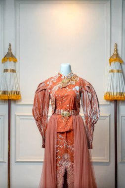 Şık bir terracotta uzun kollu gelinlik mankeni, çiçekli nakışla süslenmiş, altın kemer detaylarıyla süslenmiş ve klasik bir iç dekorla süslenmiş..