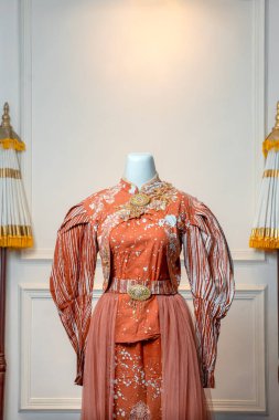 Mankenler üzerinde zarif bir terracotta gelinliği, kabarık uzun kollu, çiçekli süslemeler, altın kemer ve klasik ve dekoratif bir arka planda sergilenmiş,.