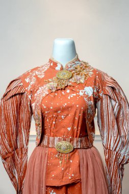 Beyaz desenli terracotta batik kumaş, kabarık uzun kollu, altın çiçekli broş, süslü kemer ve karmaşık dekoratif detaylar içeren zarif geleneksel gelinlik..