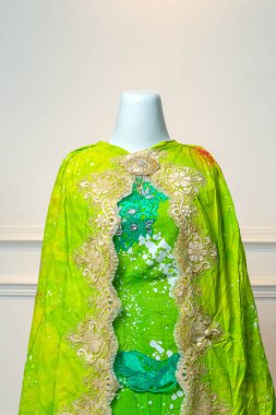 Süslü, altın işlemeli yeşil bir gelinlik, bir mankenin üzerinde sergileniyor, lüks, kültürel güzellik ve zamansız stil yayıyor..