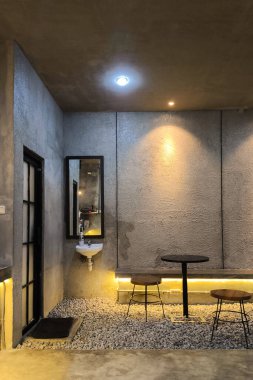 Dokulu beton duvarlar, minimalist ışıklandırma, duvara monte edilmiş lavabo, yuvarlak masa ve beyaz çakıl döşeme üzerinde tabureler içeren modern endüstriyel iç plan. Şık ve ham ortam..