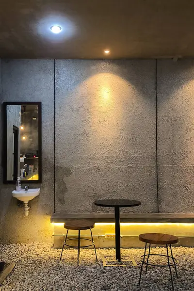 Minimalist endüstriyel arka plan desenli beton bir duvar, yuvarlak masa, iki tabure, duvara monte edilmiş lavabo, ayna, çakıl döşeme ve sıcak aksan aydınlatma. Kentsel ve şık konsept.