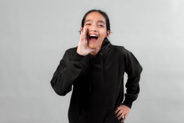 Siyah kapüşonlu bir adam elleri ağzında bağırıyor, gülümsüyor ve heyecanlı görünüyor. Stüdyoda gri bir zemine karşı duruyor..