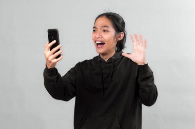 Siyah kapüşonlu heyecanlı Asyalı genç adam telefonuna şaşkınlık ve neşeyle bakıyor. Geniş gözleri ve kaldırdığı el internette beklenmedik güzel haberler aldığını gösteriyor..