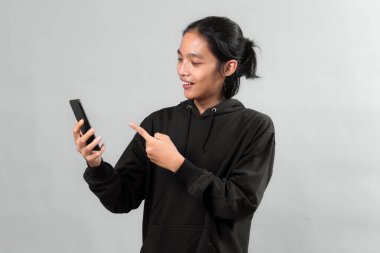 Siyah kapüşonlu uzun saçlı gülümseyen Asyalı genç bir adam bir elinde akıllı bir telefon, diğer elinde gri arka planda izole edilmiş bir şekilde ekranı işaret ediyor..