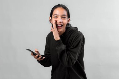 Siyah kapüşonlu heyecanlı genç Asyalı adam akıllı telefon tutuyor ve kısık sesle kameraya neşeli bir ifadeyle bakıyor, sırrı ya da flaş haber kavramını paylaşıyor..