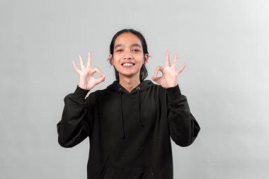 Siyah kapüşonlu gülümseyen genç Asyalı çift tamam el hareketleri sergiliyor, gri bir arka plana karşı güvenle duruyor, pozitifliğini, onayını ve memnuniyetini ifade ediyor..