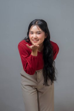 Endonezyalı kadın neşeli bir hareketle öpüşürken gülümsüyor. Uzun kollu kırmızı bir gömlek ve bej pantolon giyiyor. Sıcak, neşe ve şefkat ifade ediyor, neşeli bir havası var..