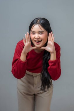 Endonezyalı kadın sanki bir şey açıklıyormuş gibi elleri ağzında gülümsüyor ve el kol hareketi yapıyor. Kırmızı uzun kollu gömlek ve bej pantolon giyiyor, neşe ve enerji saçıyor..